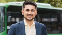 Portett av ung mann med indisk bakgrunn, smilende i dress foran grønn buss