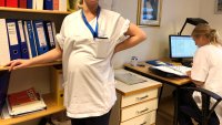 Høygravid sykepleier tar seg til ryggen og lener seg til bokhylle. Kollega sitter foran skjerm ved sine av henne.